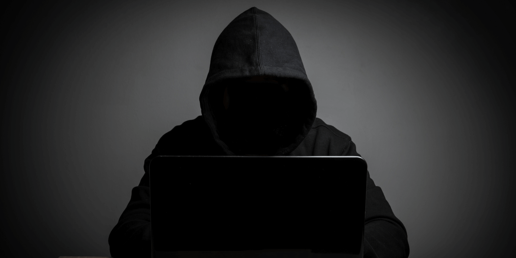 A hacker in a hoodie, looking at a laptop, obscured by shadow.