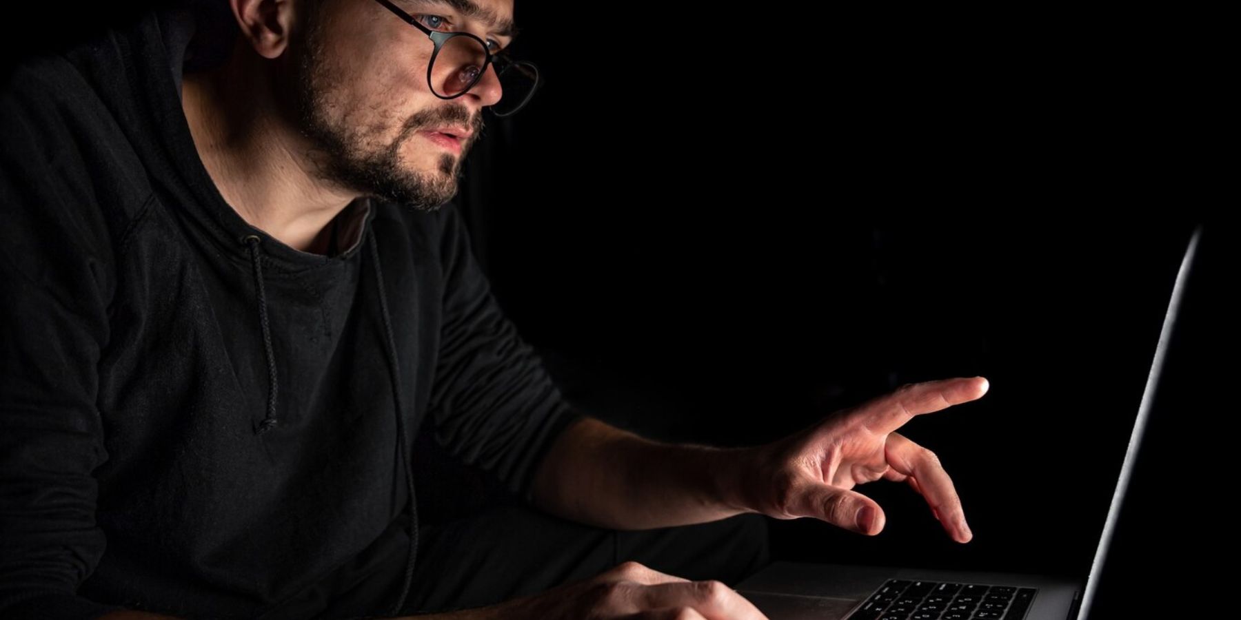 Man wearing eyeglasses in a dimly lit room, attentively pointing at his laptop screen.