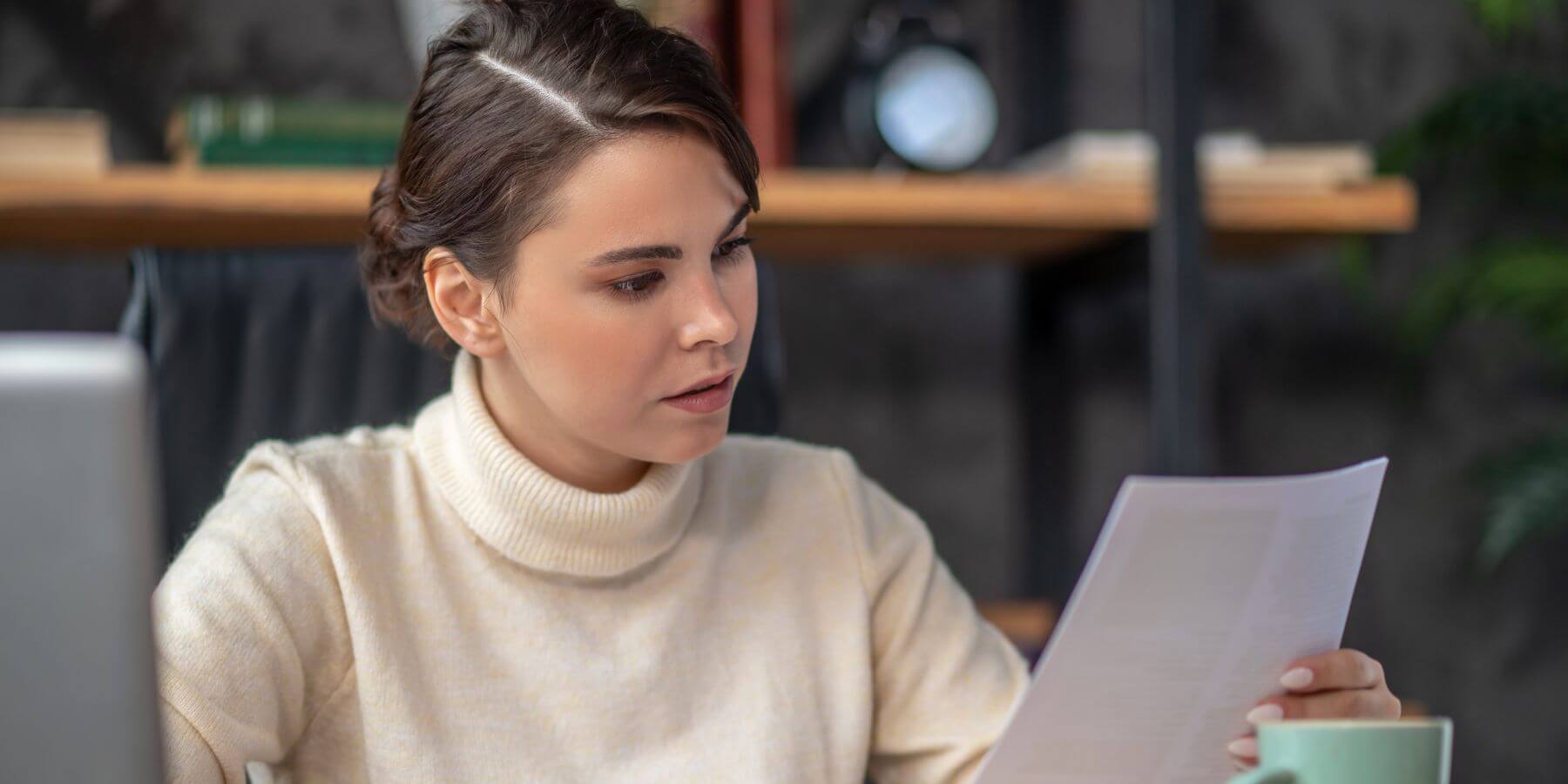 A stressed woman sitting at her office desk, intensely scrutinizing a document in her hand, with an expression of worry on her face.