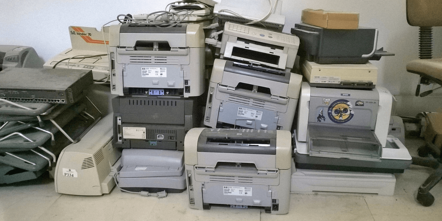 A large stack of unused printers piled haphazardly on top of each other, symbolizing excess and redundancy in office equipment.